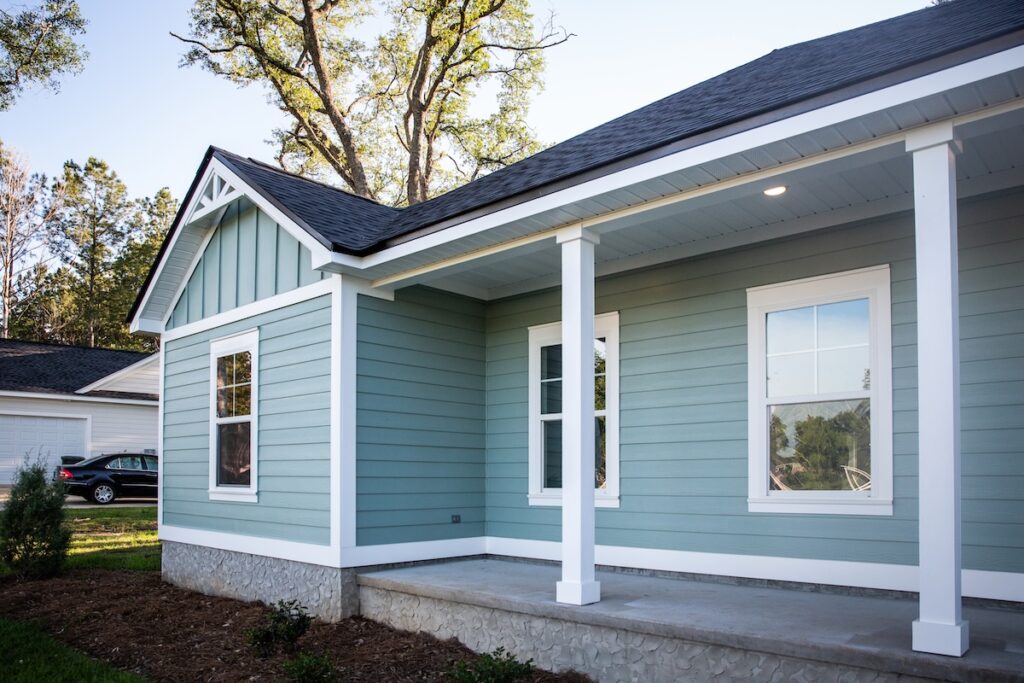 A modern light blue house with white trim, featuring large windows and a small covered front porch with white columns. Trees and a parked car are visible in the background.