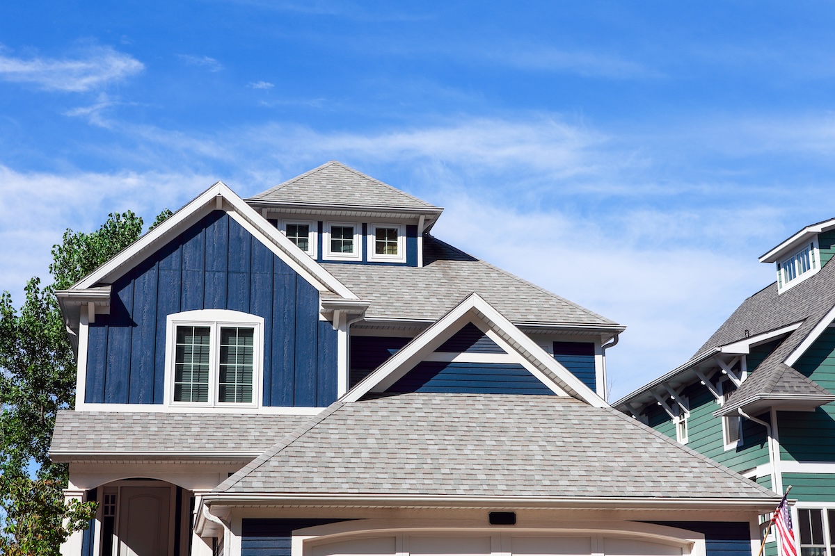 A modern house with dark blue siding, white trim, and gray shingle roof—expertly crafted by roofing East Norriton Township PA—set against a bright blue sky with wispy clouds. Neighboring green house partially visible on the right.