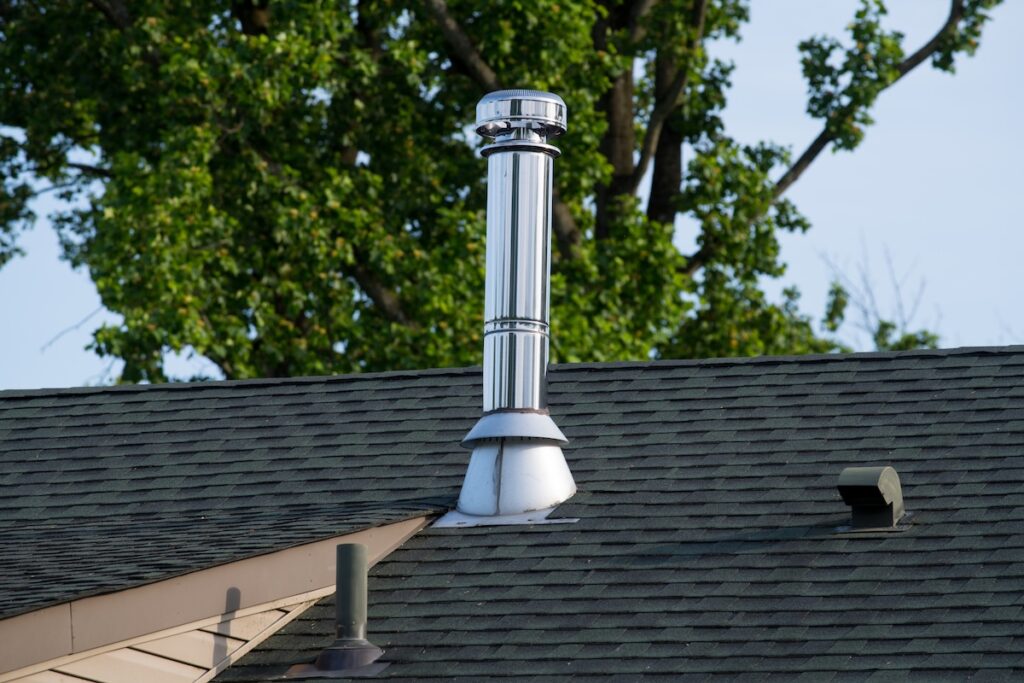A shiny metal chimney pipe extends upward from a shingled roof, with green leafy trees in the background. The roof has dark shingles and a vent near the chimney.