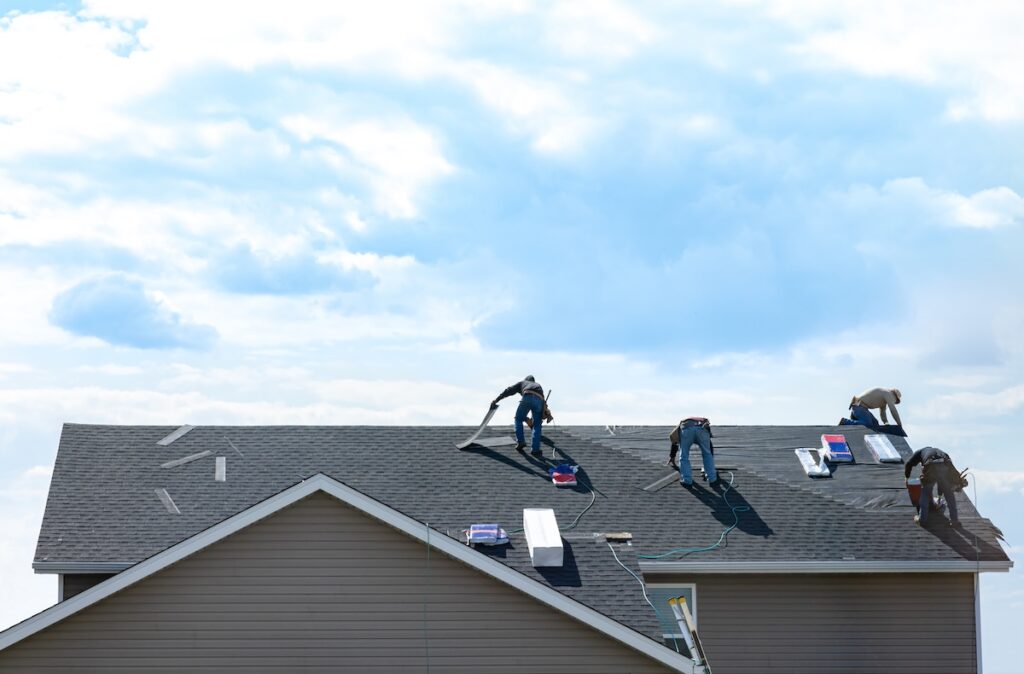 Four workers are installing or repairing shingles on the roof of a house under a partly cloudy sky, with roofing materials and tools spread around them.