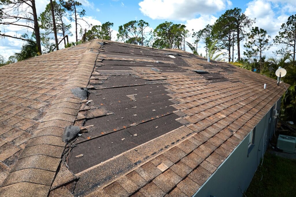 A house roof with many missing and damaged shingles exposes the black underlayment, raising the question: how often should you replace your roof? Loose and askew shingles suggest it may be time. Trees and a blue sky with clouds are in the background.