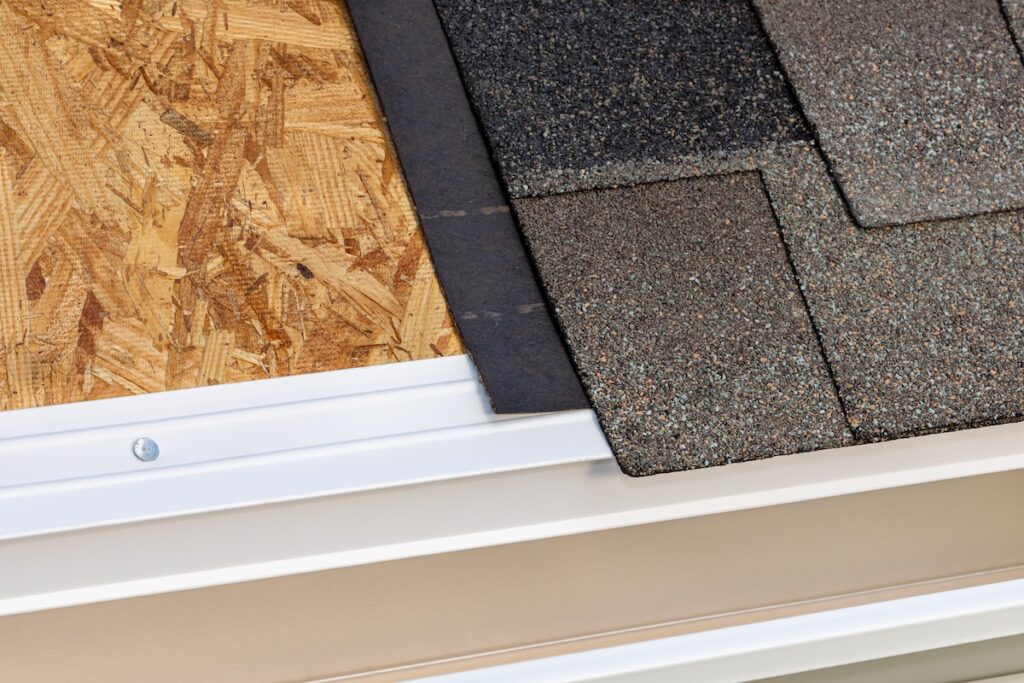 Close-up of a roof edge showing layers: a sheet of oriented strand board (OSB), roofing felt, and asphalt shingles, next to a white metal drip edge above a beige gutter.