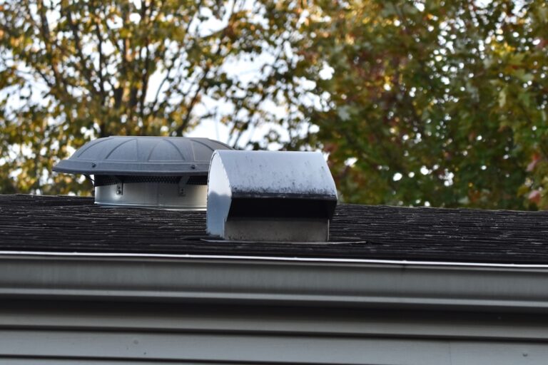 Two metal roof vents are mounted on a dark shingle roof, with green trees in the blurred background. One vent is round with a wide top, and the other is rectangular with a slanted cover.