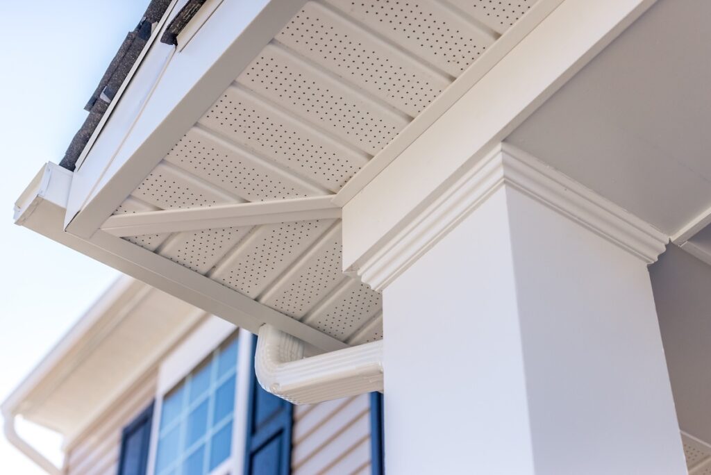 Close-up view of a house’s exterior showing a white column, soffit, and gutter system beneath the roof, with part of a window and blue sky visible in the background.