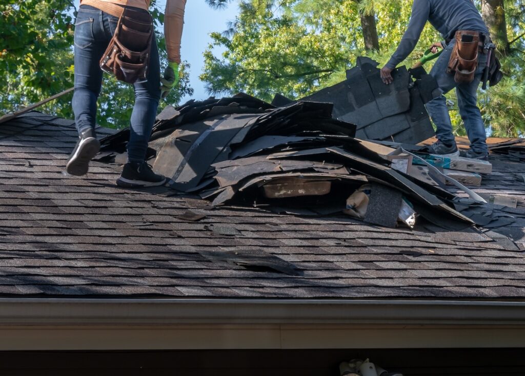 Two workers in jeans and tool belts are removing old shingles from a house roof on a sunny day. Piles of discarded roofing materials are scattered on the roof beneath their feet. Trees with green leaves are visible in the background.