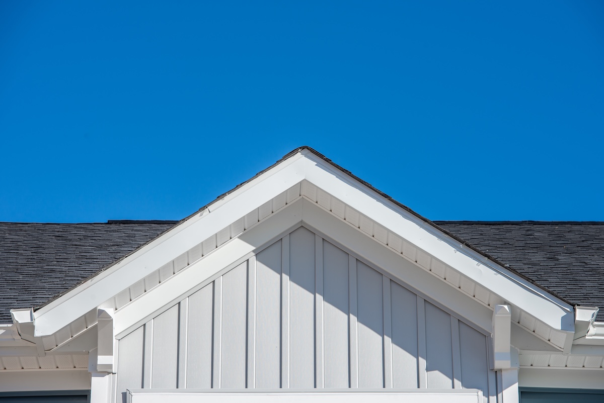 The image shows the peak of a modern house roof with white trim and light siding against a clear blue sky. The roof has dark shingles and the architecture features clean, sharp lines.